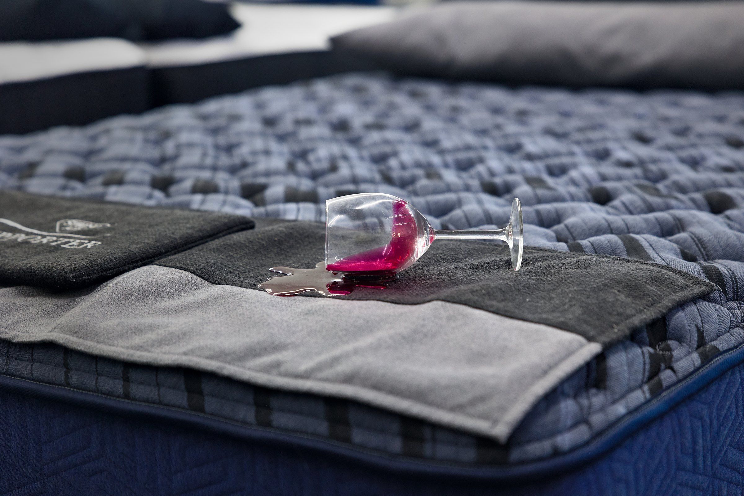 A tipped-over wine glass spilling on a mattress protector, the wine beading on top