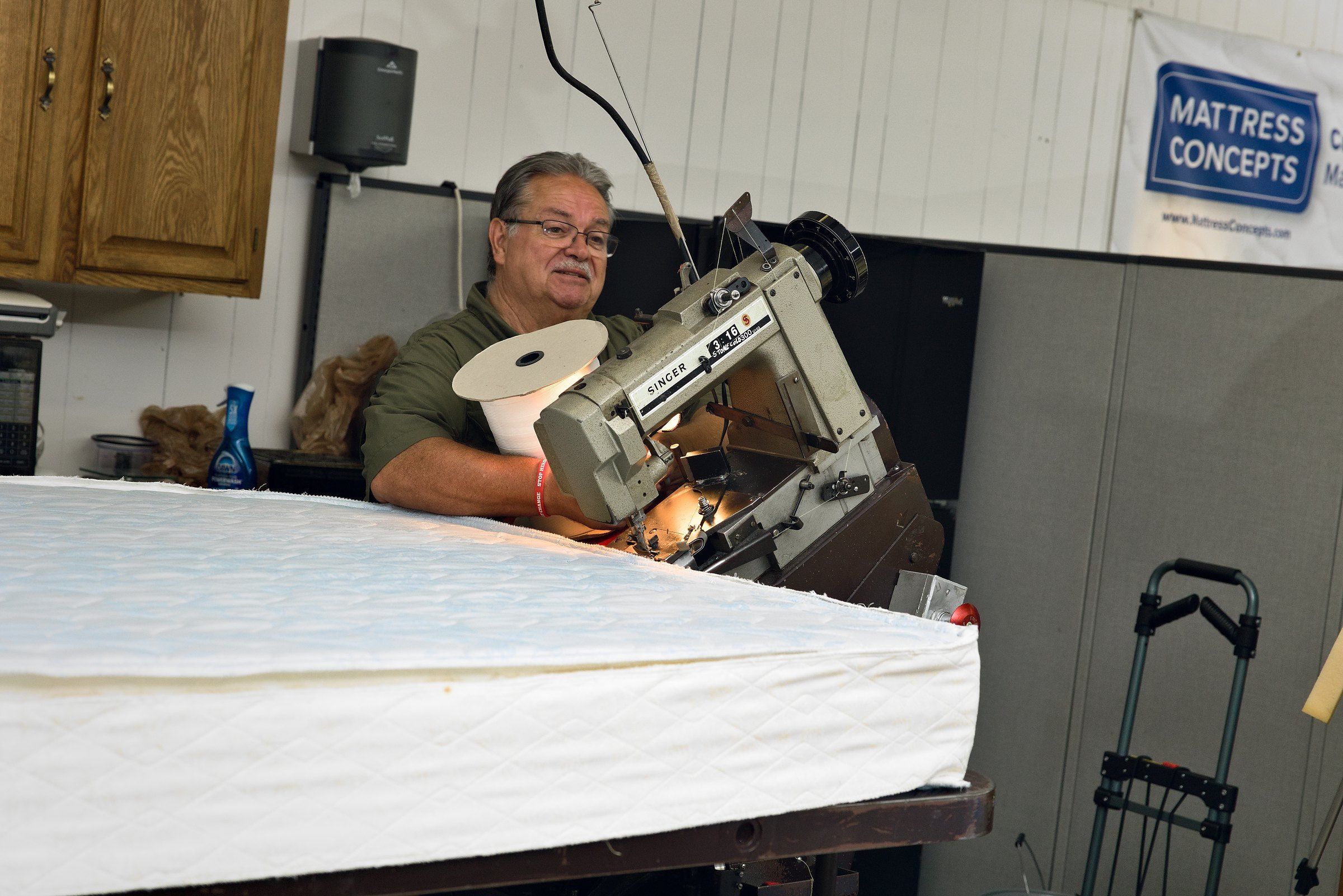 Clinton smiling behind the tape-edge machine, with the Mattress Concepts banner on the wall