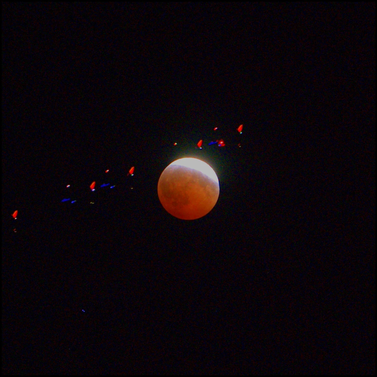 A copper eclipsed moon with a diagonal trail of red and white aircraft navigation lights running up across it from lower left to upper right.