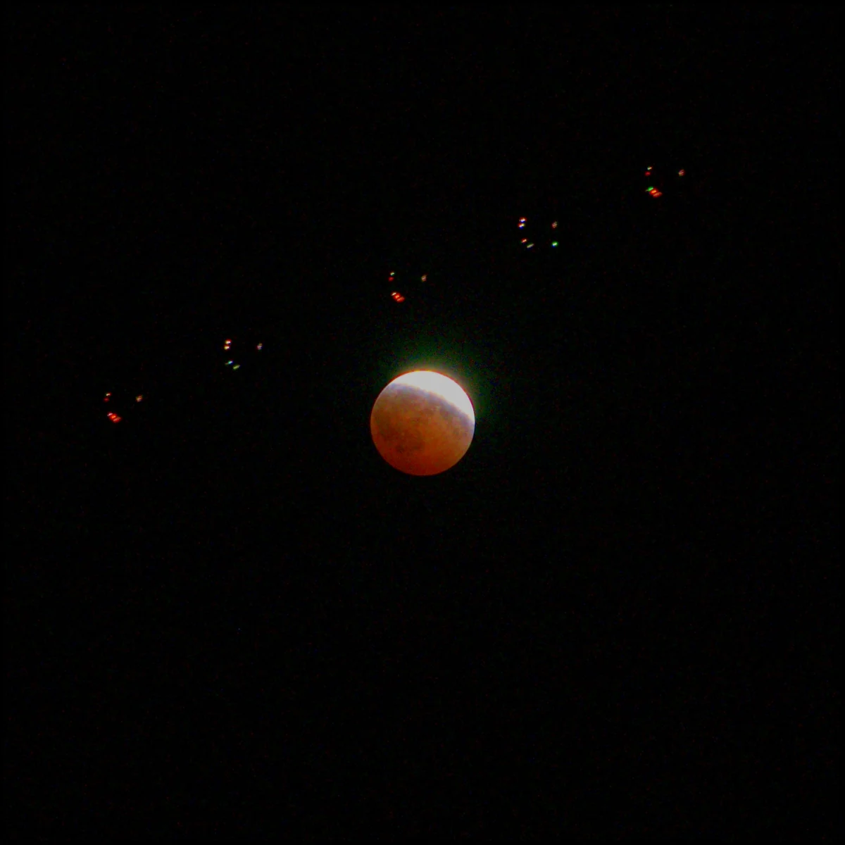 The same eclipsed moon lower in the frame, with a line of coloured navigation lights arcing above and to the left of it.