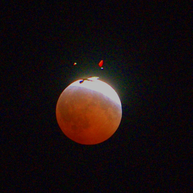 An airliner in silhouette against the bright edge of a partly eclipsed copper moon, its navigation lights showing as red and white dashes above.