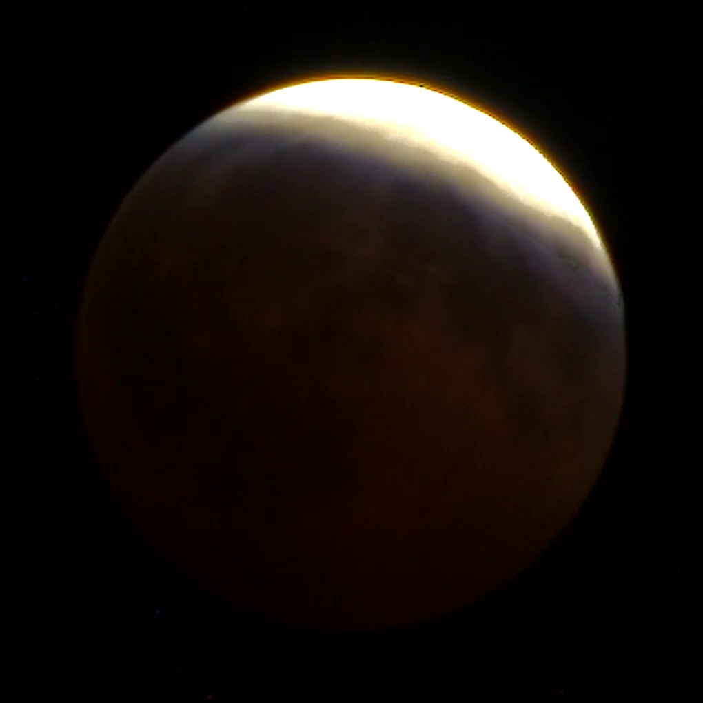 The partly eclipsed moon with a brilliant white and gold rim along the upper right edge and the rest of the disc a dim copper, with maria visible through the shadow.