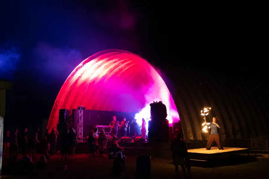 A quonset stage lit deep pink at night, a fire performer out front.
