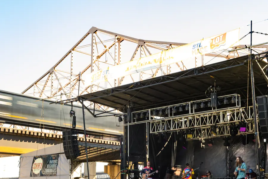 An outdoor stage set under a steel bridge in daylight.